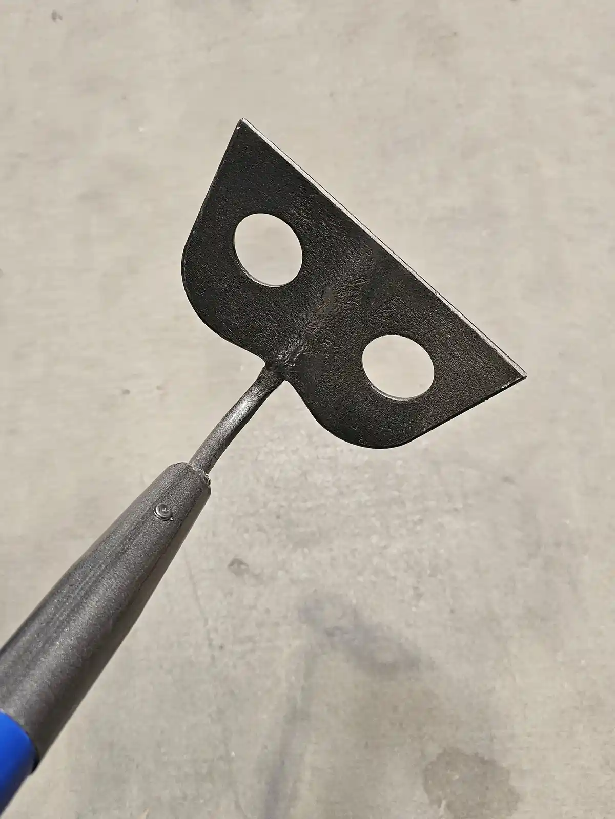A close-up photo shows a metal garden tool with a blue handle and a black, triangular head featuring two circular holes. The…
