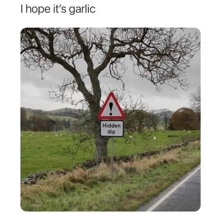 A roadside warning sign in a rural area, featuring a red triangle with an exclamation mark above a white rectangle labeled…