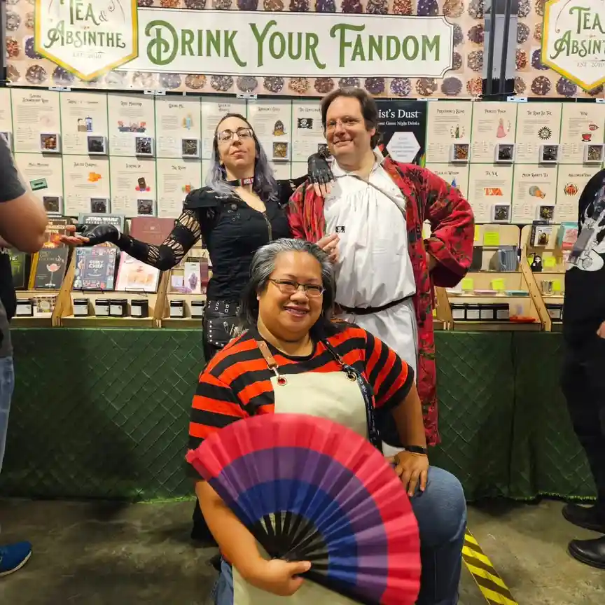 Three people pose at a 'Tea & Absinthe' booth with a banner reading 'DRINK YOUR FANDOM' behind them. The person in front…