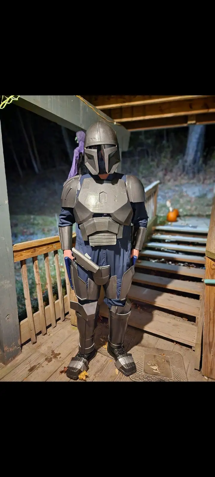 A person in a full Mandalorian armor costume stands on a wooden porch at night, holding a blaster. The costume includes a…
