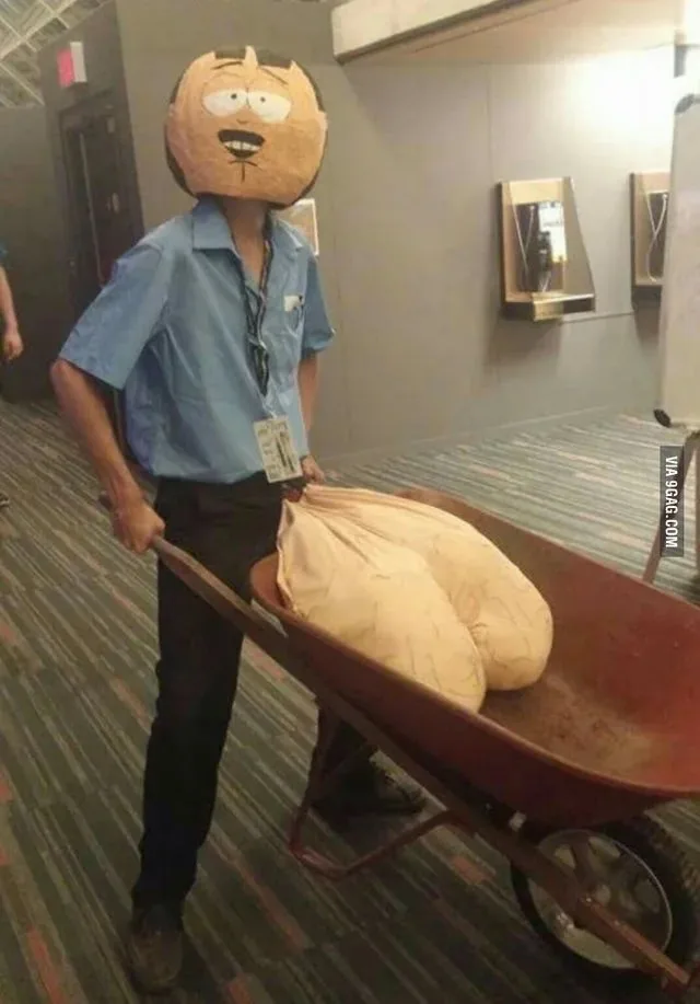 A person wearing a large, cartoonish head mask of a character with a wide grin and thick eyebrows pushes a red wheelbarrow…