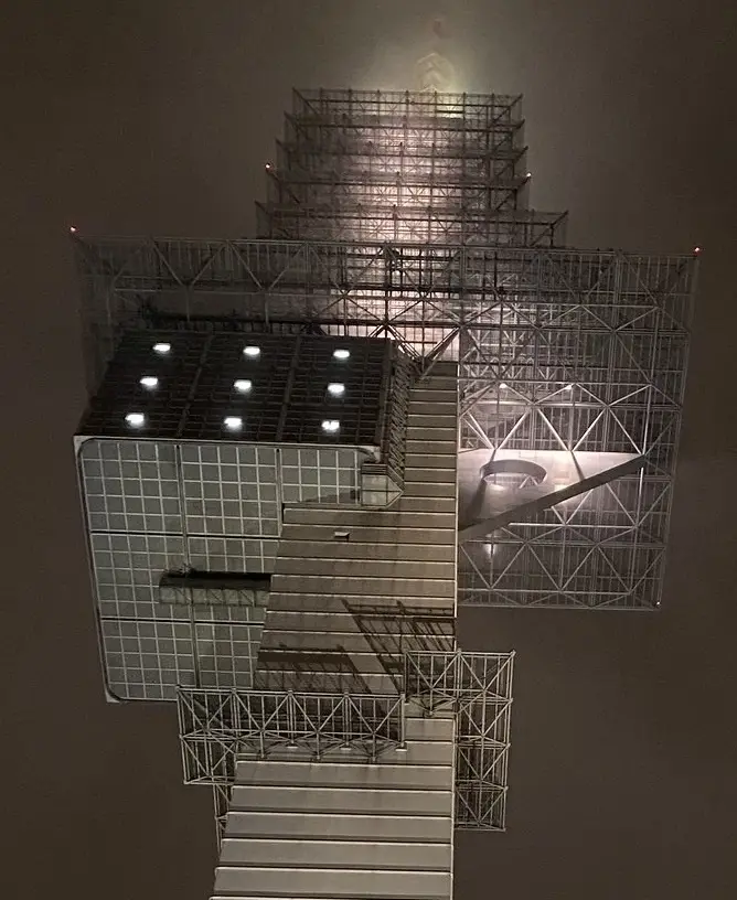 A photograph of a large, intricate metal scaffolding structure resembling a staircase or tower, illuminated from within…