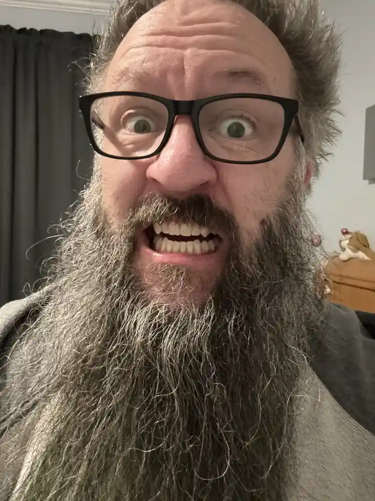 A close-up selfie of a man with a long gray beard, wearing black-rimmed glasses, making an exaggerated, wide-eyed, toothy…