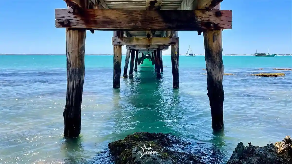 A scenic photo of a weathered wooden pier extending over clear turquoise water under a bright blue sky, with sailboats…