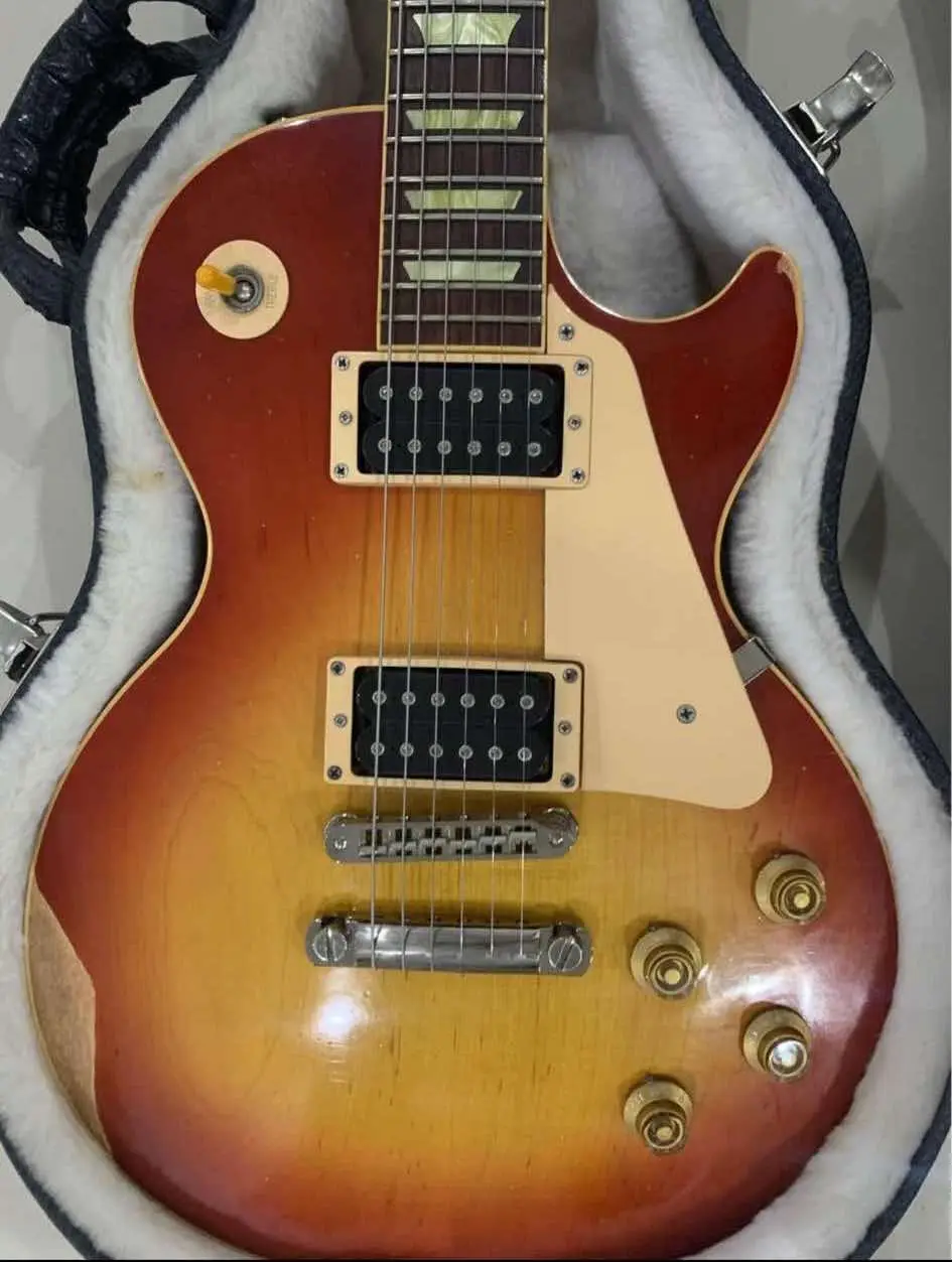A sunburst-finish electric guitar, resembling a Gibson Les Paul, is shown resting inside its plush-lined case. The guitar's…