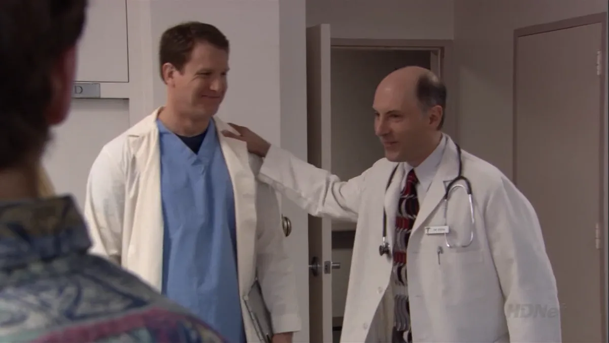 Two men in white doctor's coats stand in a hospital hallway. The man on the right, bald and wearing a stethoscope, has his…