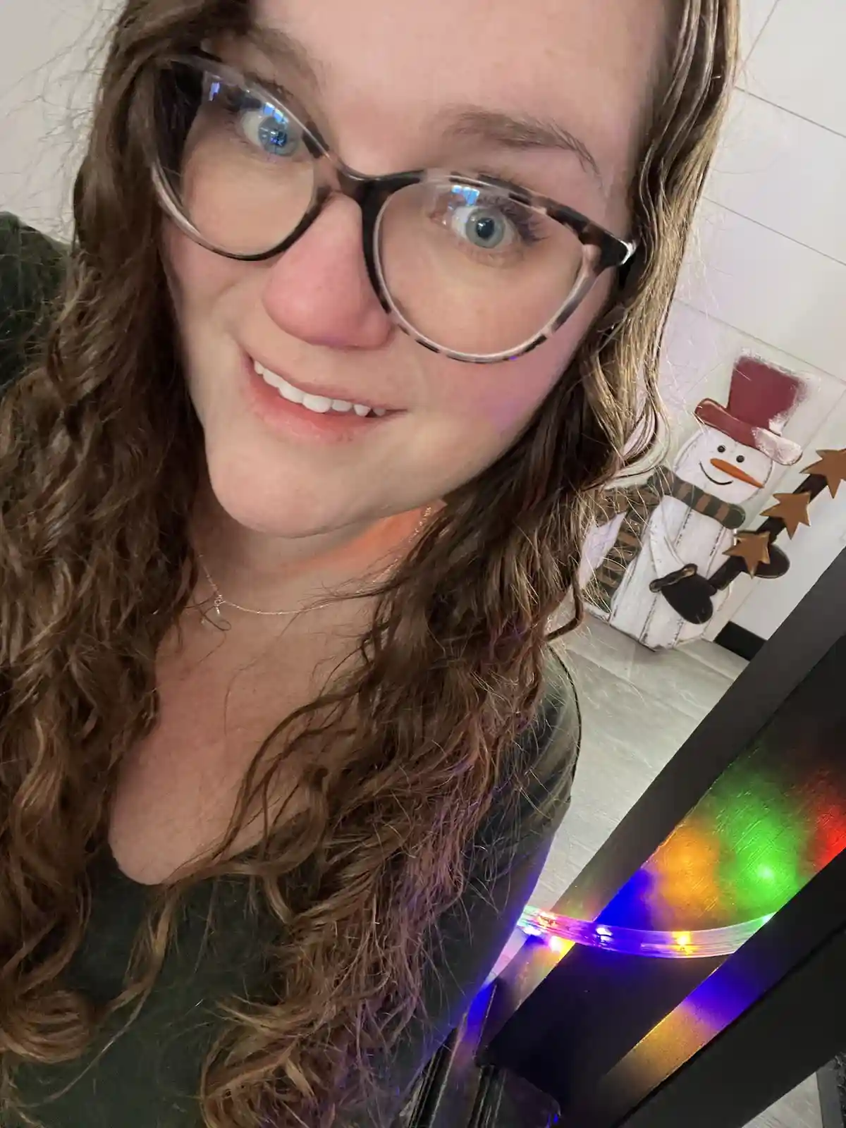 A selfie of a woman with glasses and long brown hair, smiling with wide eyes, standing near a snowman decoration and…