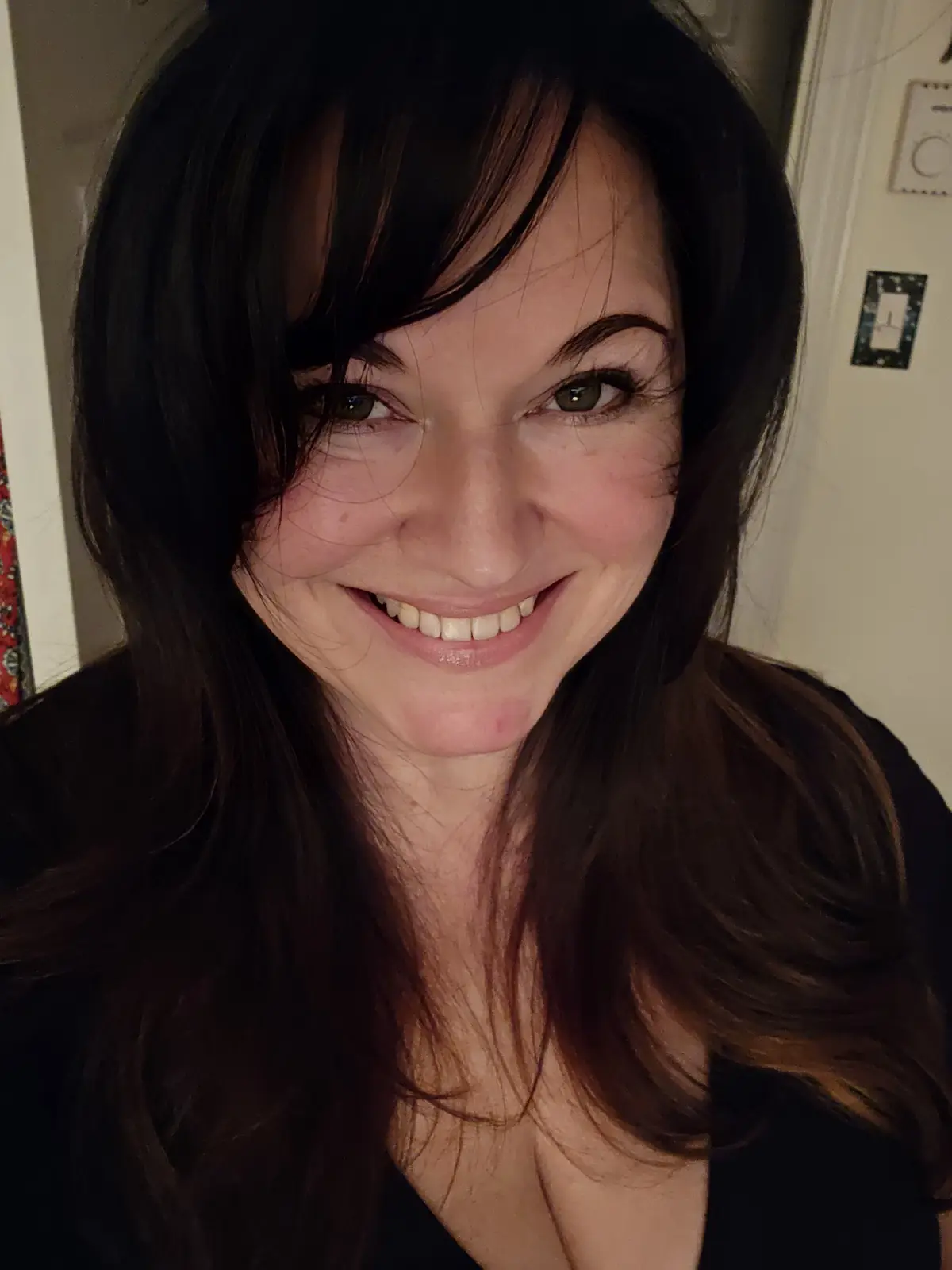 A close-up selfie of a woman with dark, shoulder-length hair and a wide, toothy smile, looking directly at the camera. She…