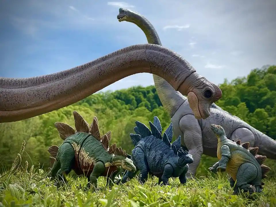 Three small Stegosaurus toy dinosaurs stand in a grassy field, looking up at a giant Brachiosaurus toy dinosaur whose long…