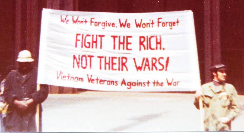 Two men hold a large white banner with red text that reads 'We Won't Forgive, We Won't Forget FIGHT THE RICH. NOT THEIR…