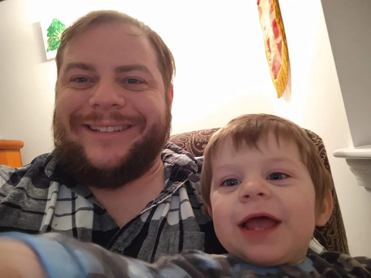 A man with a beard and a young child are smiling at the camera in a selfie. The man is wearing a plaid shirt and the child…