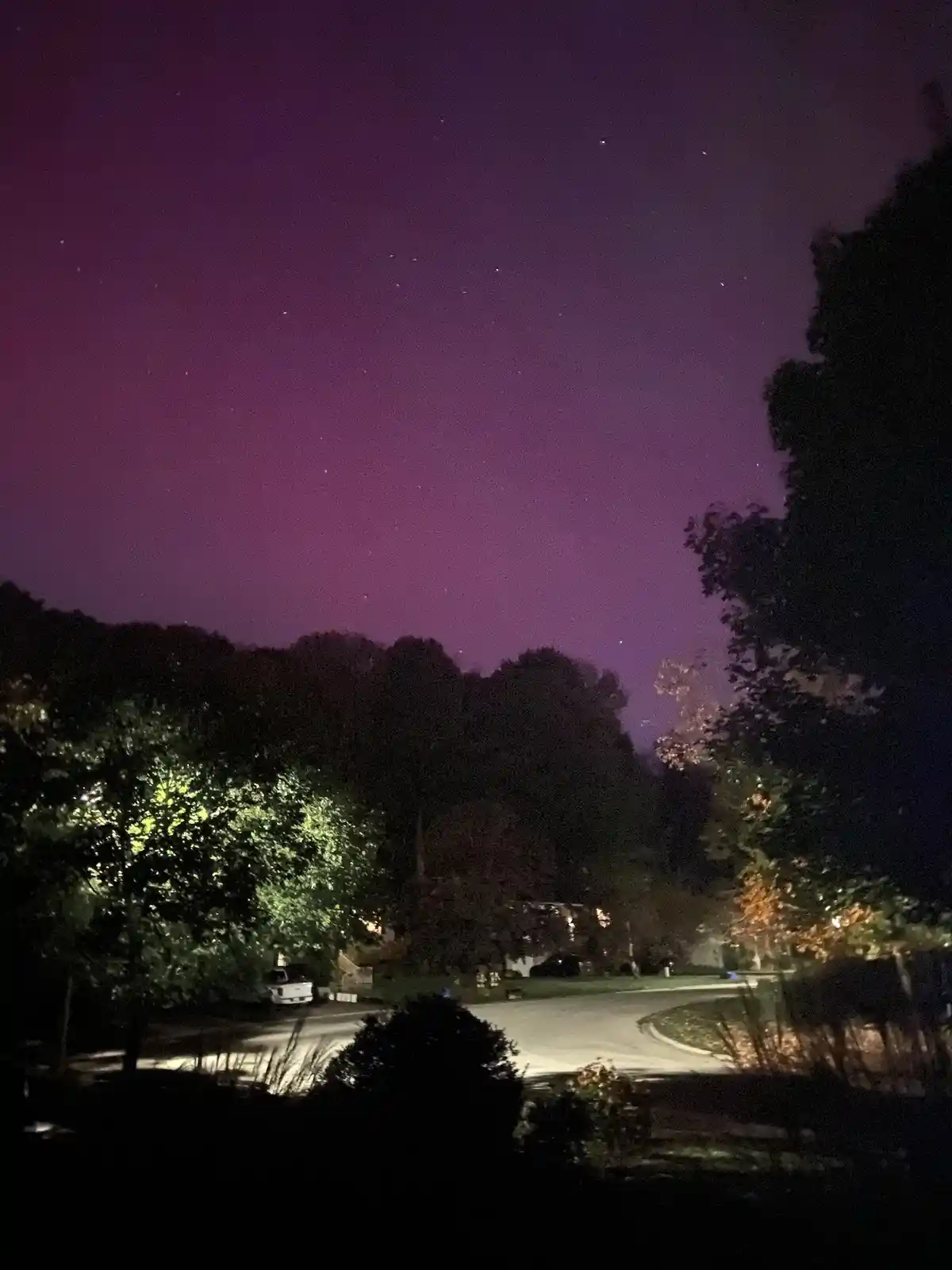 A nighttime photo shows a dark suburban street with trees and a white truck, under a purple-hued sky filled with visible…