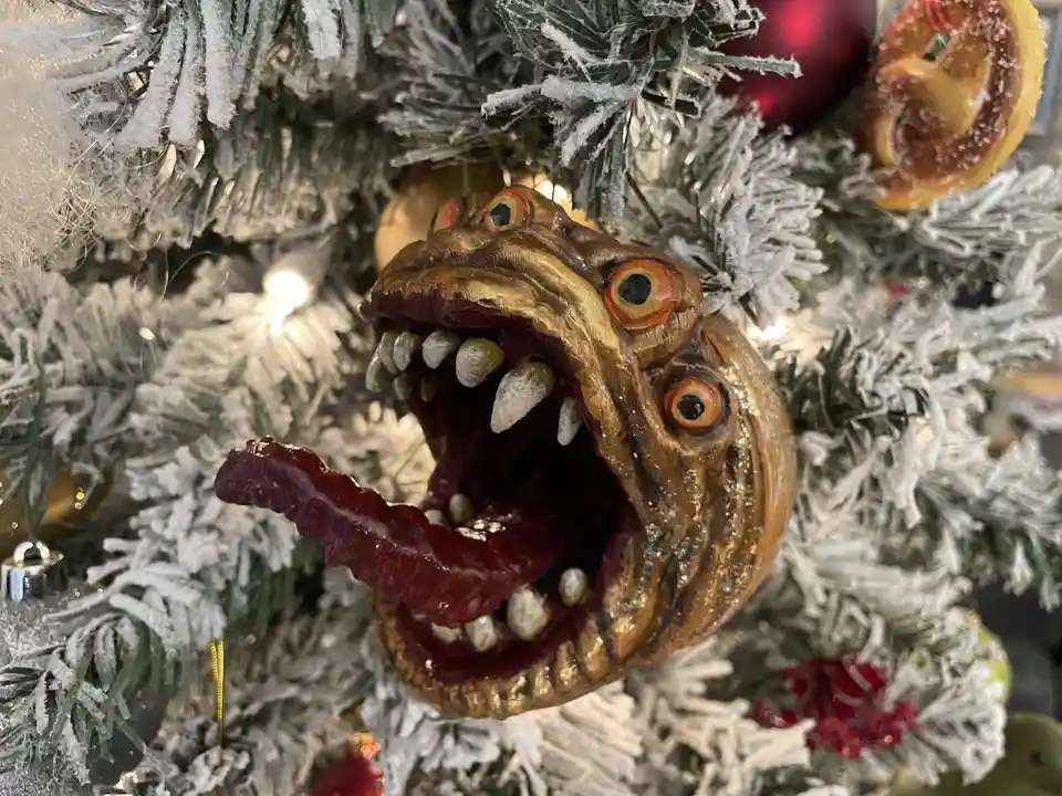 A close-up photo shows a grotesque, gilded monster ornament with three bulging orange eyes, sharp white teeth, and a long…