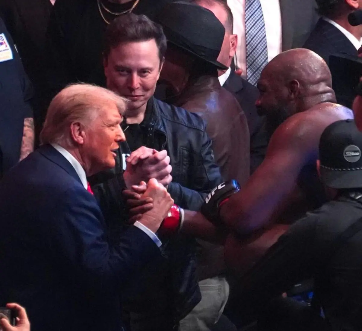A photo shows Donald Trump arm wrestling a shirtless, muscular man while Elon Musk watches with a smirk. The overlaid text…
