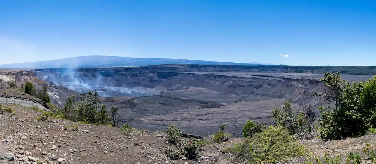 A panoramic view of a vast, steaming volcanic crater under a clear blue sky, with rugged, rocky terrain and sparse green…