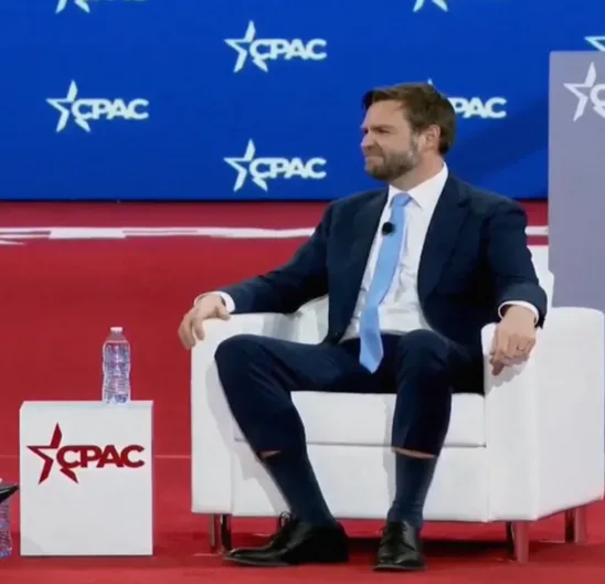 a man in a suit sits in a white chair on a red carpet stage with a CPAC logo backdrop. A water bottle sits on a CPAC-branded…
