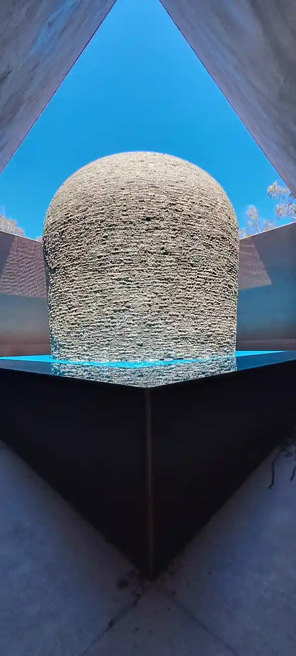 A low-angle photo shows a large, textured, dome-shaped stone sculpture sitting in a shallow pool of bright blue water…