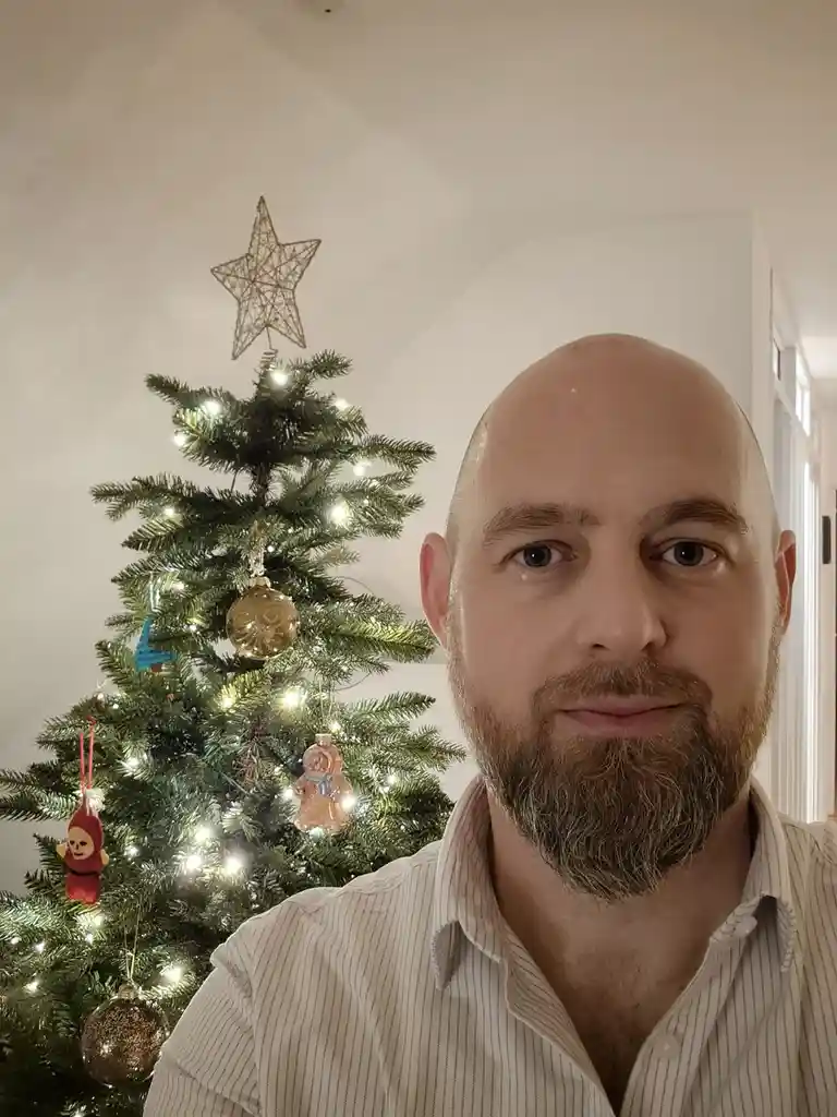 A bald man with a beard in a striped shirt looks directly at the camera with a neutral expression, standing in front of a…
