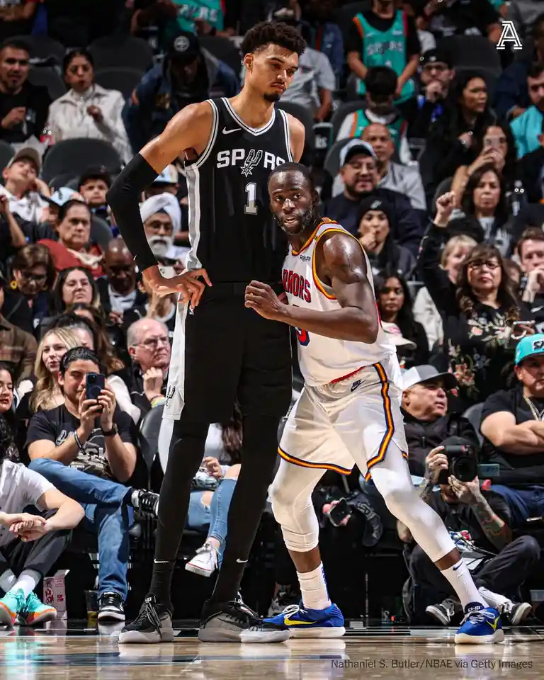 A basketball player in a white Warriors jersey is looking up at a much taller Spurs player in a black jersey with the number…