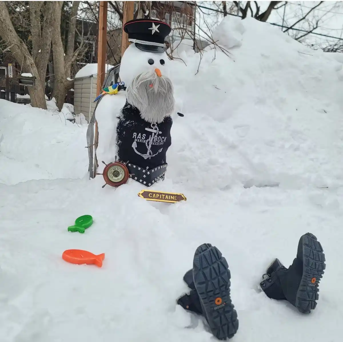 A snowman dressed as a captain with a hat, beard, and vest featuring an anchor and text, sits in a snowy yard with a wooden…