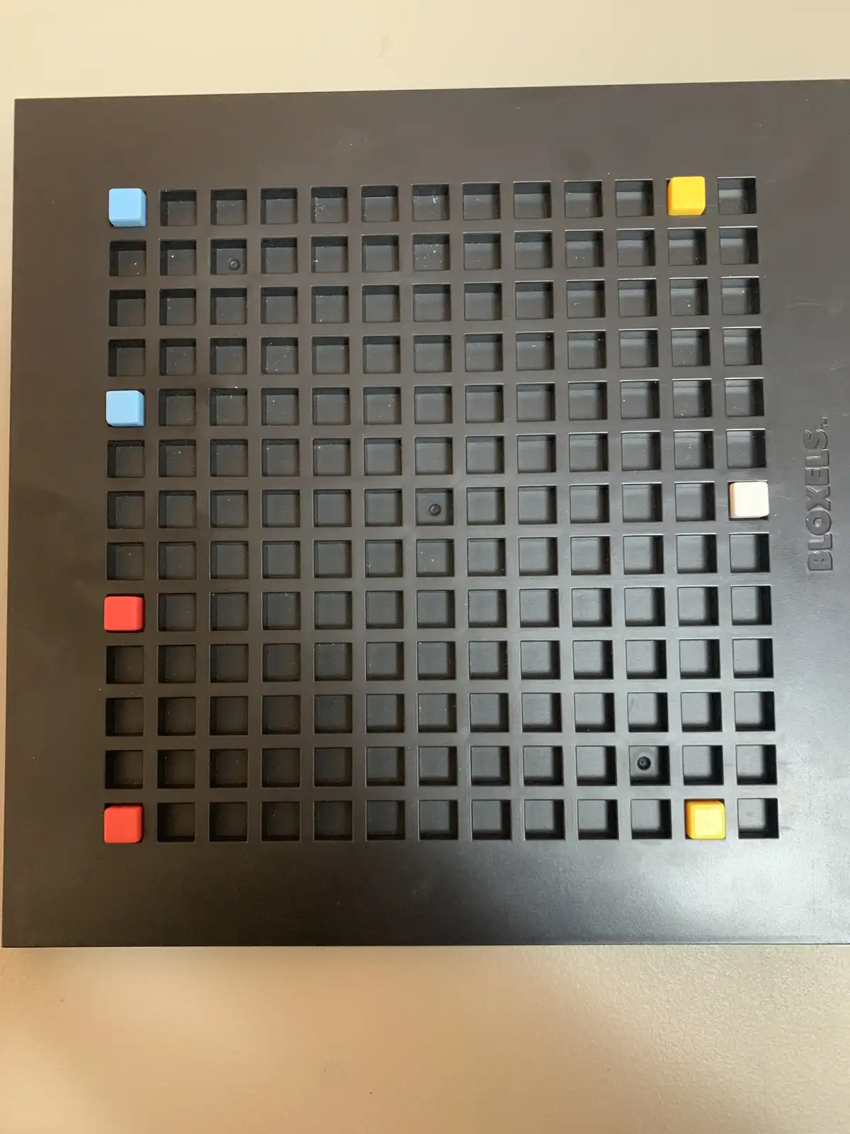 A black Bloxels grid board with a few colorful square tiles placed in specific holes, including blue, red, yellow, and white…