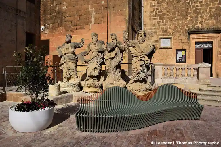 Four weathered stone statues of bearded men in robes stand on a raised platform behind a modern, wavy, green metal bench…