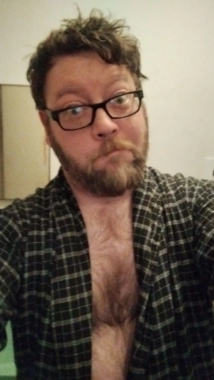 A man with glasses and a beard stares at the camera with a confused or exaggerated expression, wearing an open plaid shirt…