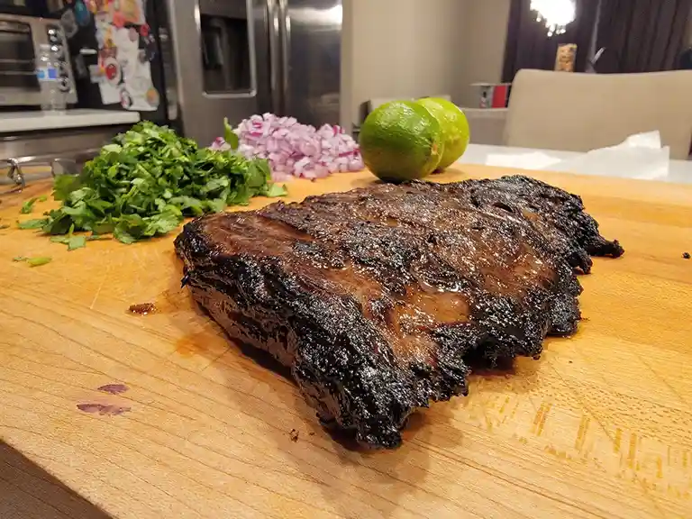 A large, heavily charred piece of meat sits on a wooden cutting board, surrounded by chopped cilantro, diced red onion, and…