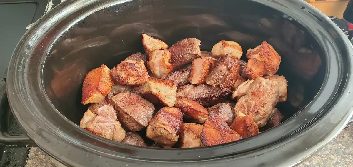 A black ceramic slow cooker is filled with browned, seared chunks of meat, likely beef or pork, showing a crispy exterior…