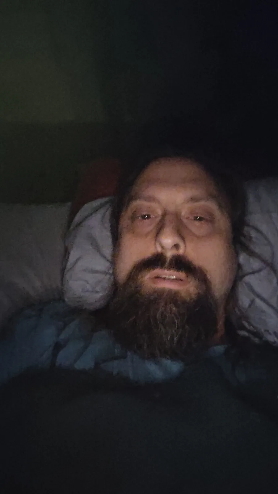 A man with a full beard lies in bed in a dimly lit room, looking directly at the camera with a weary expression. He is…