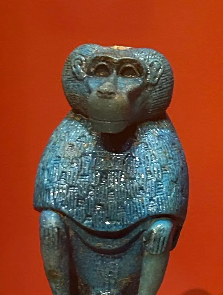 A blue, ancient Egyptian statue of a monkey, covered in hieroglyphs, stands against a solid red background. The overlaid…