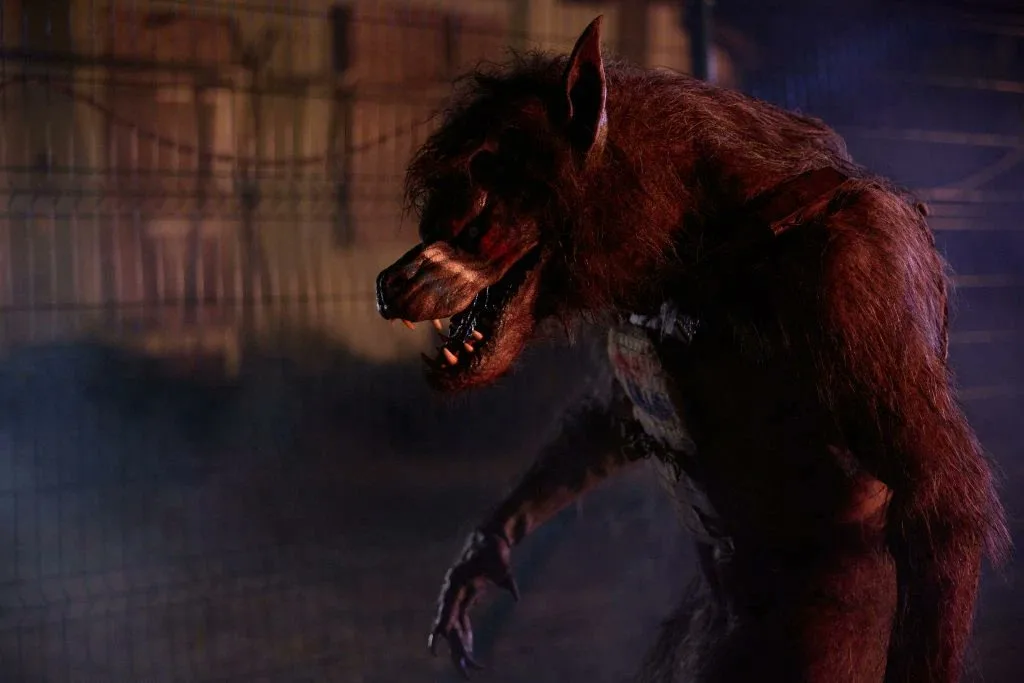 A menacing, hairy werewolf with bared teeth and glowing eyes stands in a dimly lit, smoky enclosure with a chain-link fence…