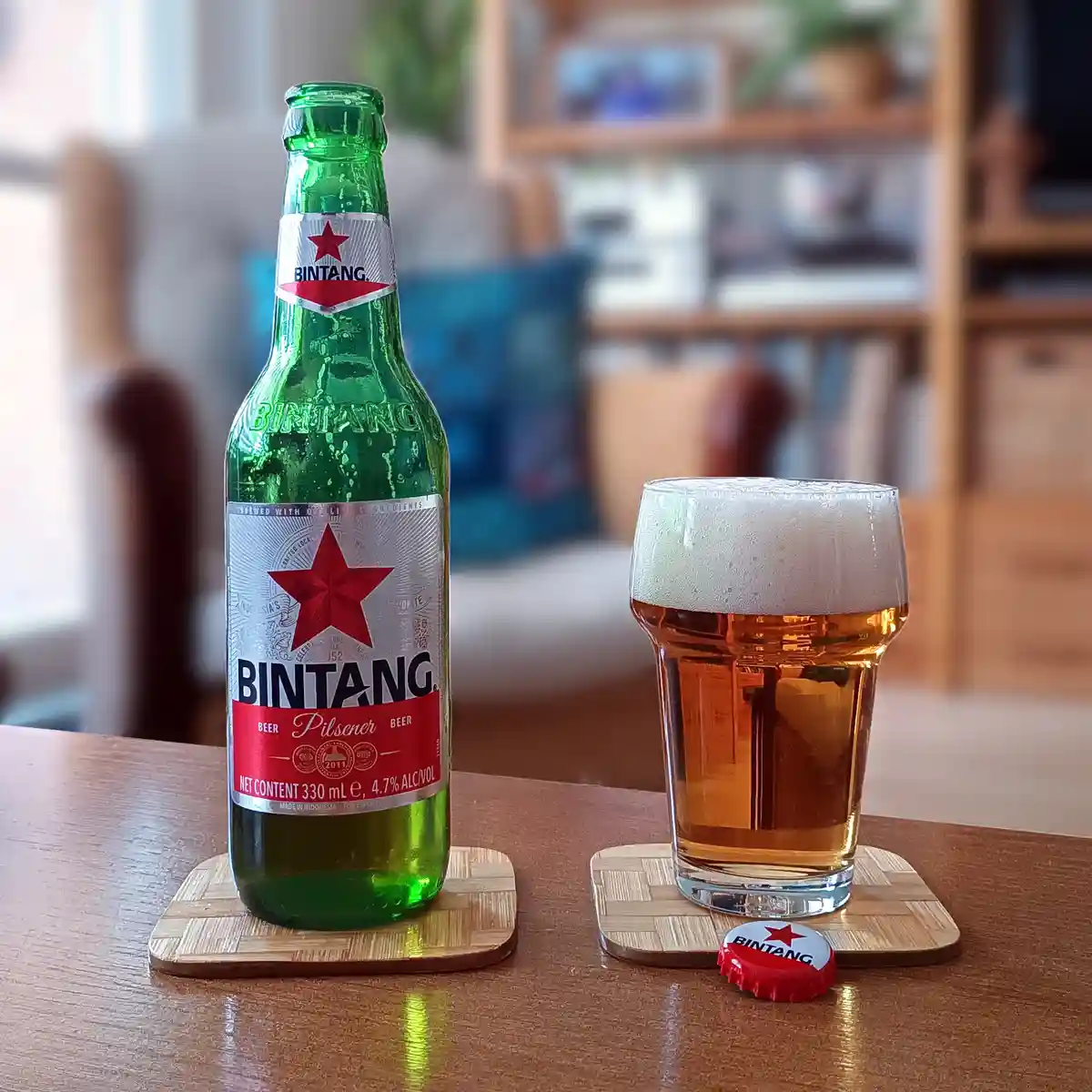 A green Bintang beer bottle and a full glass of beer sit on wooden coasters on a table, with the red Bintang bottle cap…