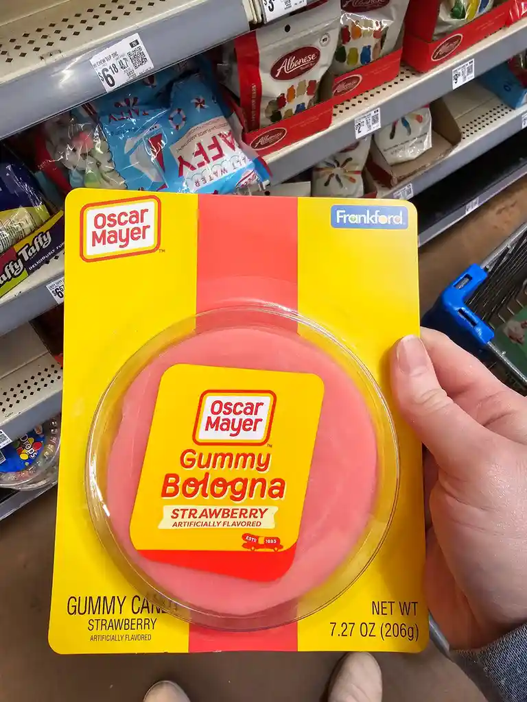 A hand holds a yellow and red package of Oscar Mayer Gummy Bologna, strawberry flavored, in a grocery store aisle. The…