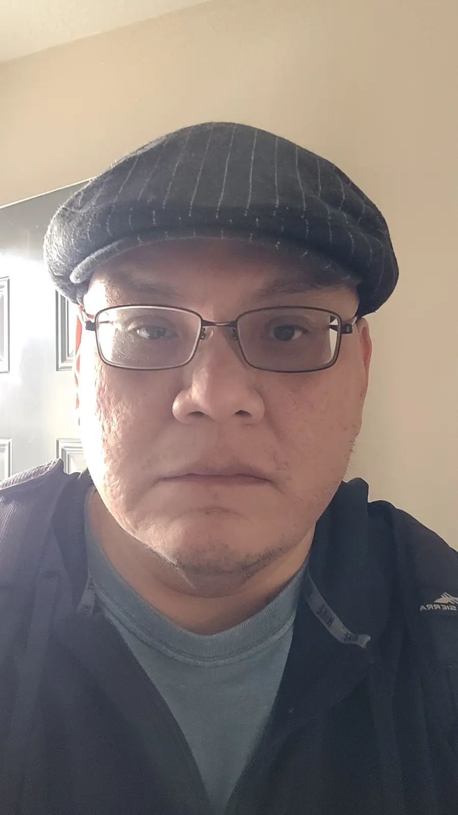 A man with glasses and a pinstripe flat cap stares directly at the camera with a deadpan, unimpressed expression. He wears a…