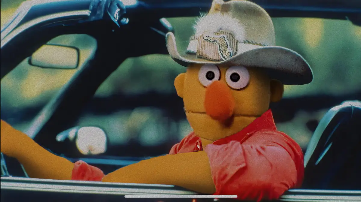A yellow Muppet with a large orange nose, wearing a cowboy hat with a crocodile patch and a red shirt, sits in the driver's…
