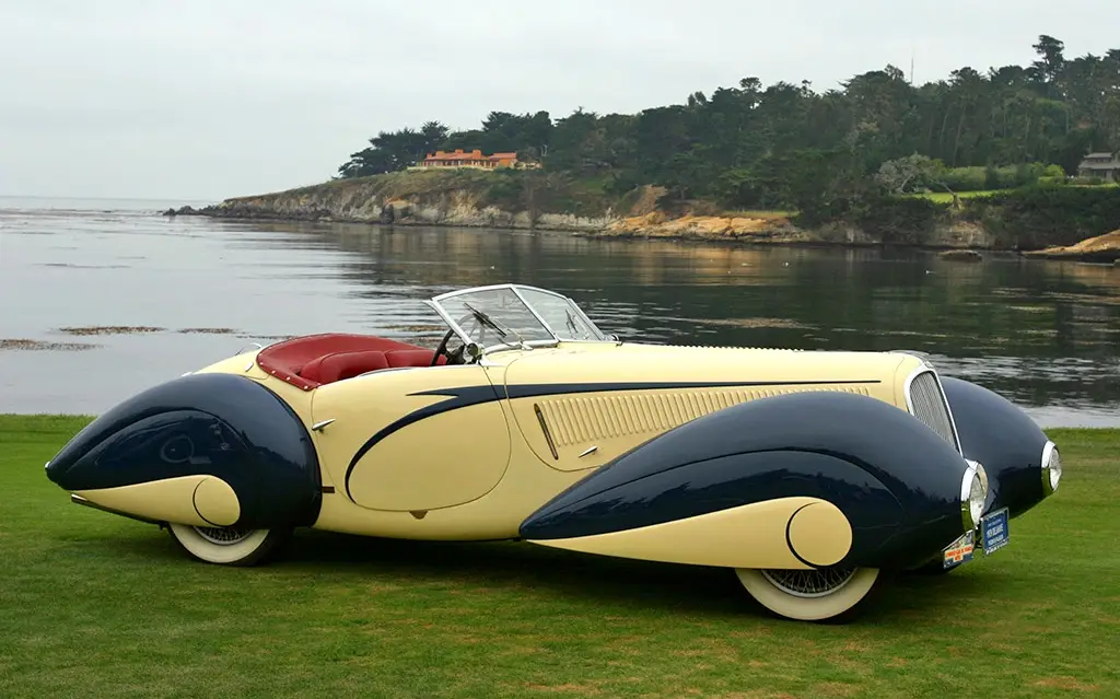 a vintage 1937 darrin sportster, a cream and navy blue convertible with a red interior, is parked on green grass beside a…