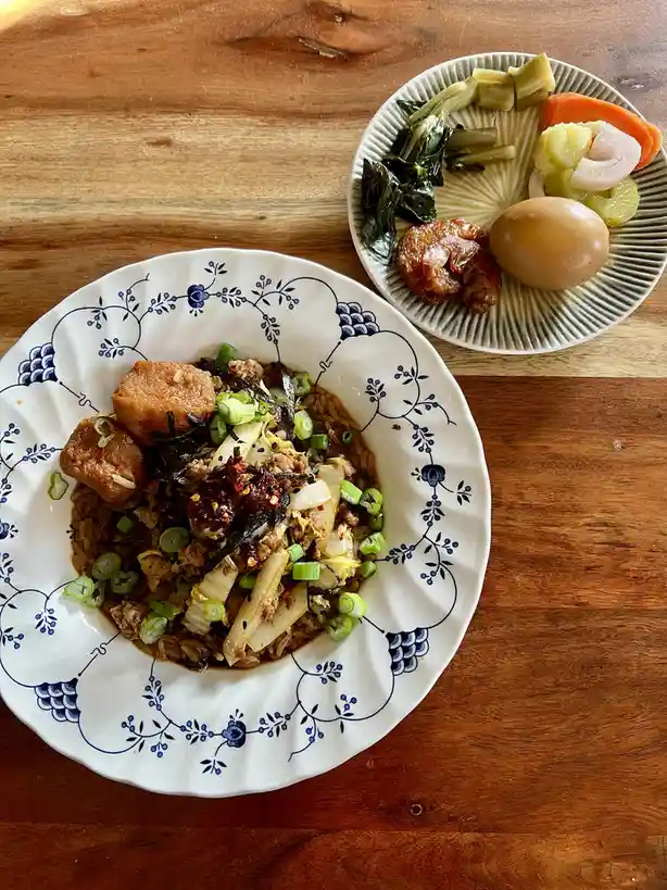 A top-down photo shows a bowl of noodle dish with tofu, green onions, and chili oil, next to a side plate with pickled…