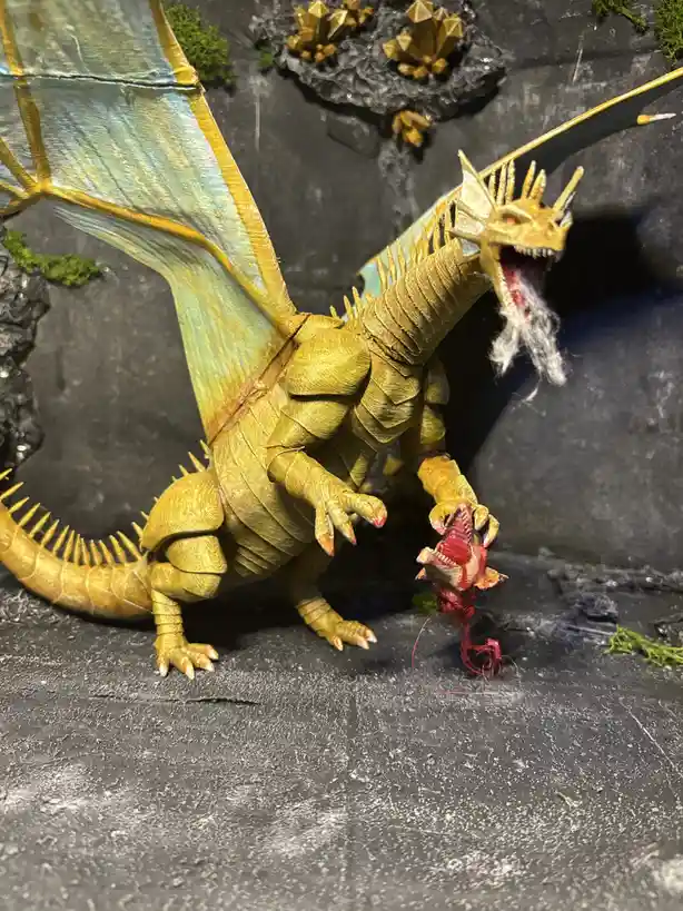 A large, golden-yellow dragon figurine with translucent blue wings and a long spiked tail stands on a dark, rocky surface…