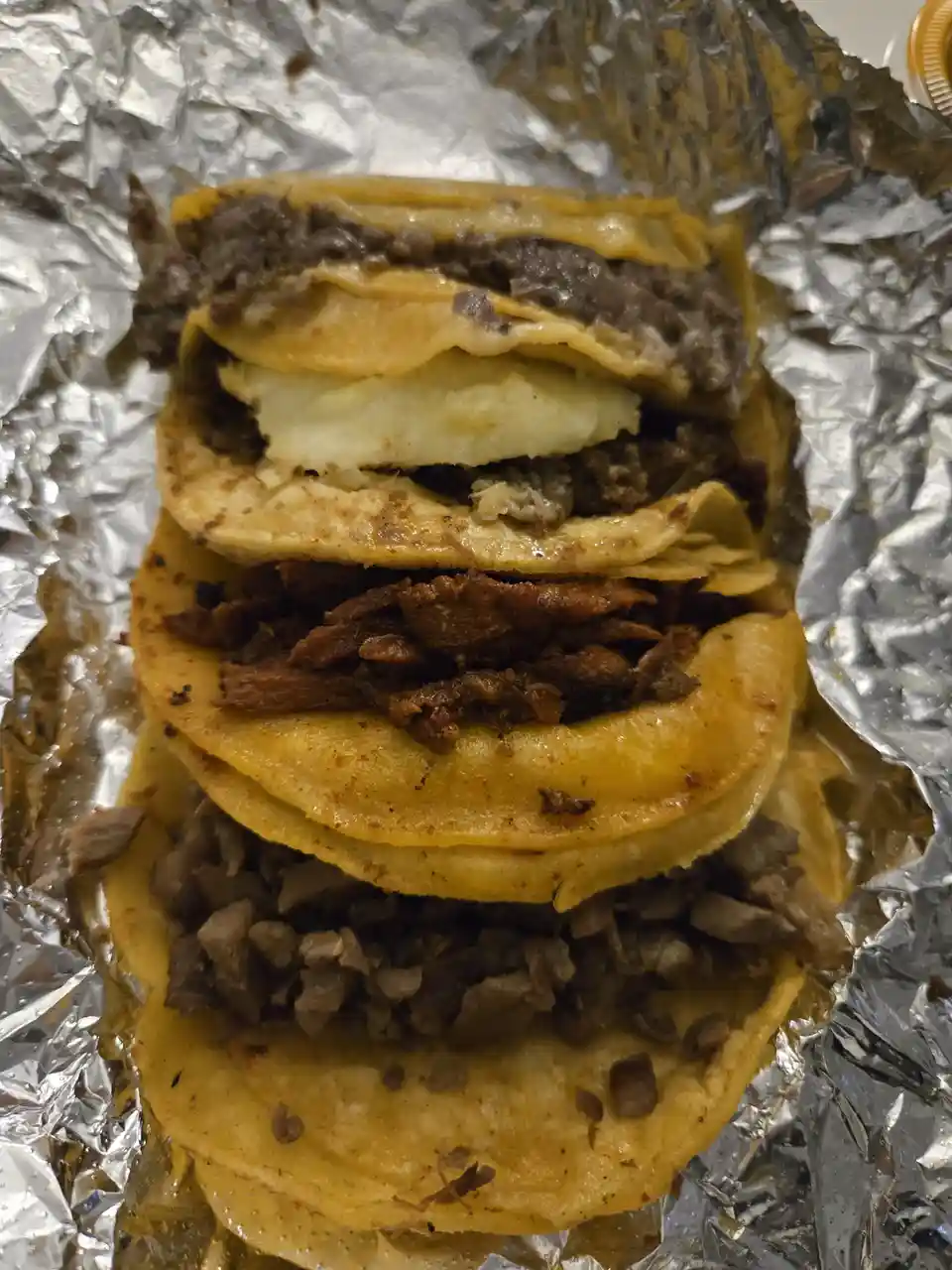 Four tacos are stacked vertically on crinkled aluminum foil, each filled with different types of meat and toppings like…