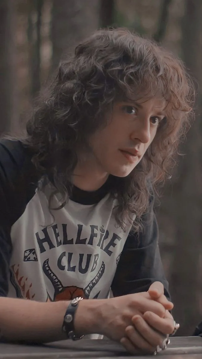 a young man with long, wavy brown hair sits at a table in a wooded area, wearing a white and black t-shirt with 'HELLFIRE…