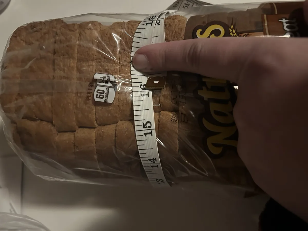 A hand holds a measuring tape wrapped around a loaf of bread in plastic packaging, with the tape showing measurements around…