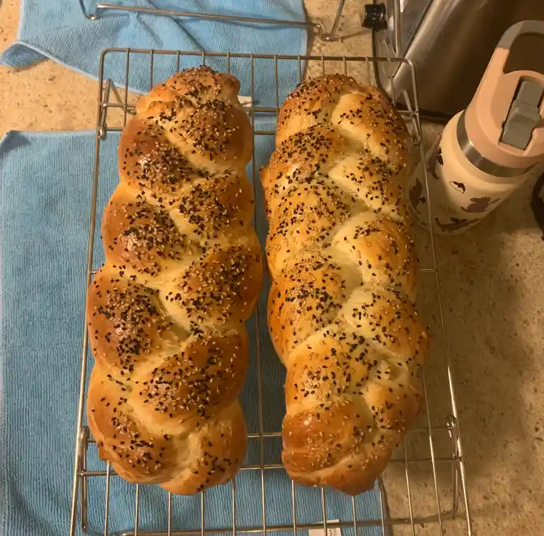 Two golden-brown braided loaves of bread, topped with black and white sesame seeds, sit side-by-side on a metal cooling rack…