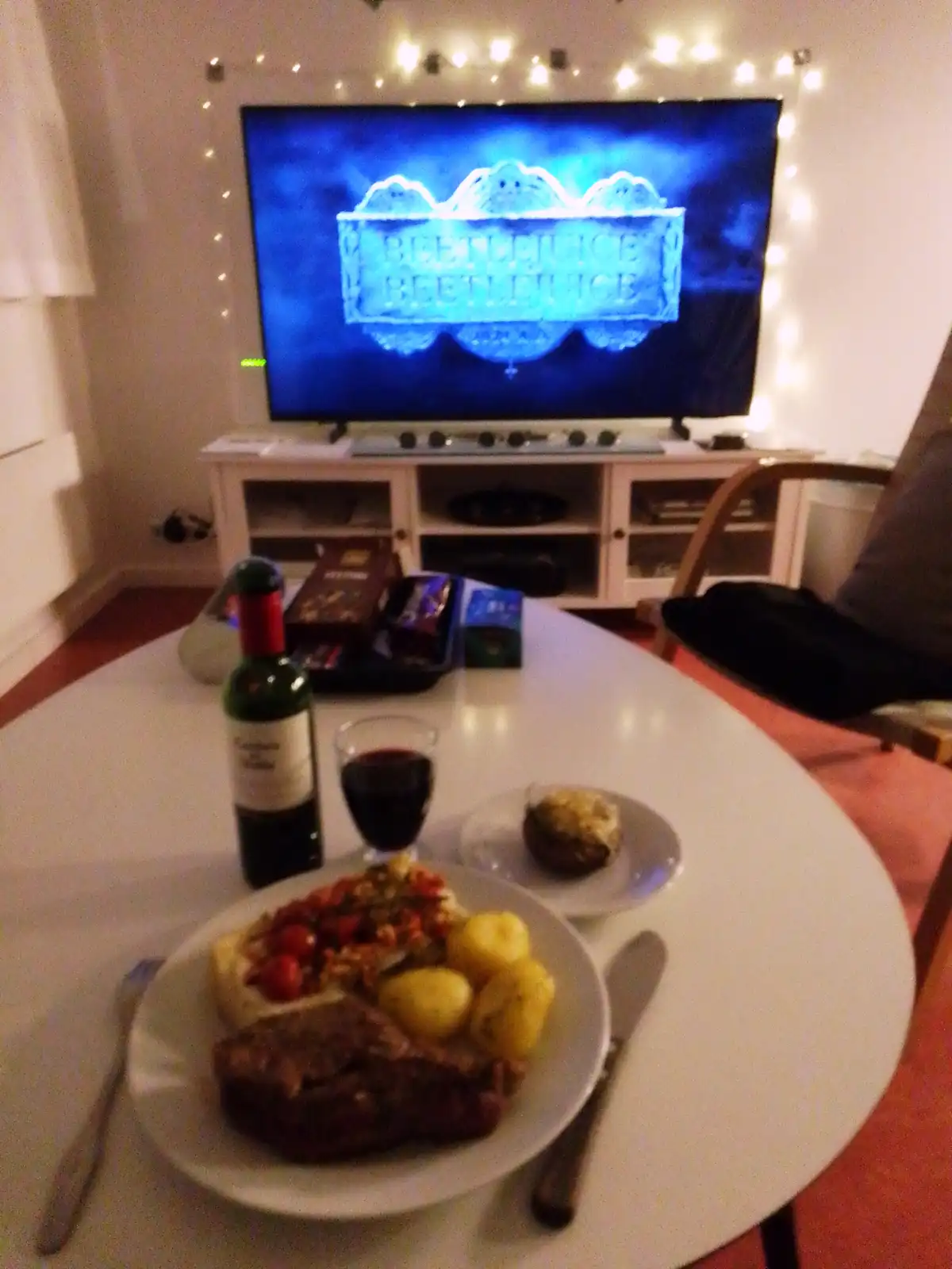 A cozy living room scene shows a white table in the foreground with a large plated meal including steak, potatoes, and…