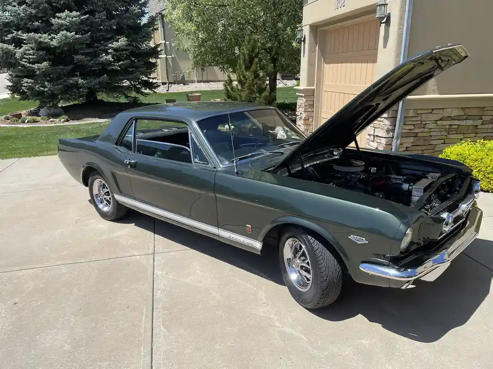 A dark green classic Ford Mustang with its hood open, parked on a concrete driveway in front of a house with a garage. The…