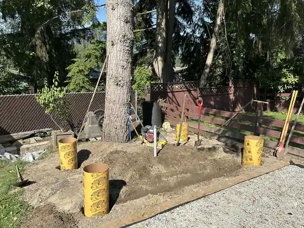 An outdoor backyard scene shows a large, freshly dug hole surrounded by yellow cardboard tubes, shovels, and other gardening…