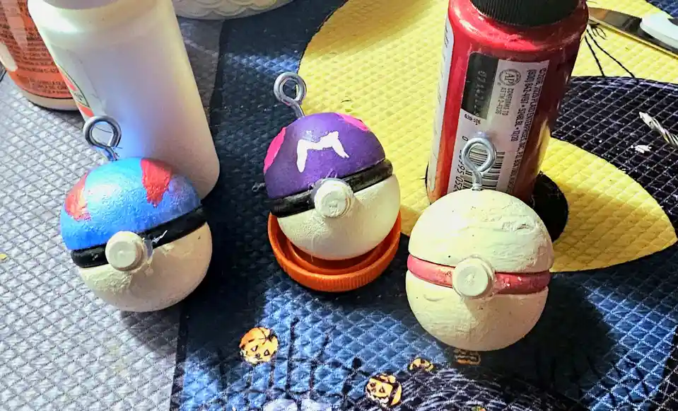 Three handmade, crudely painted Pokémon Poké Ball crafts sit on a Halloween-themed mat next to art supplies. The balls are…