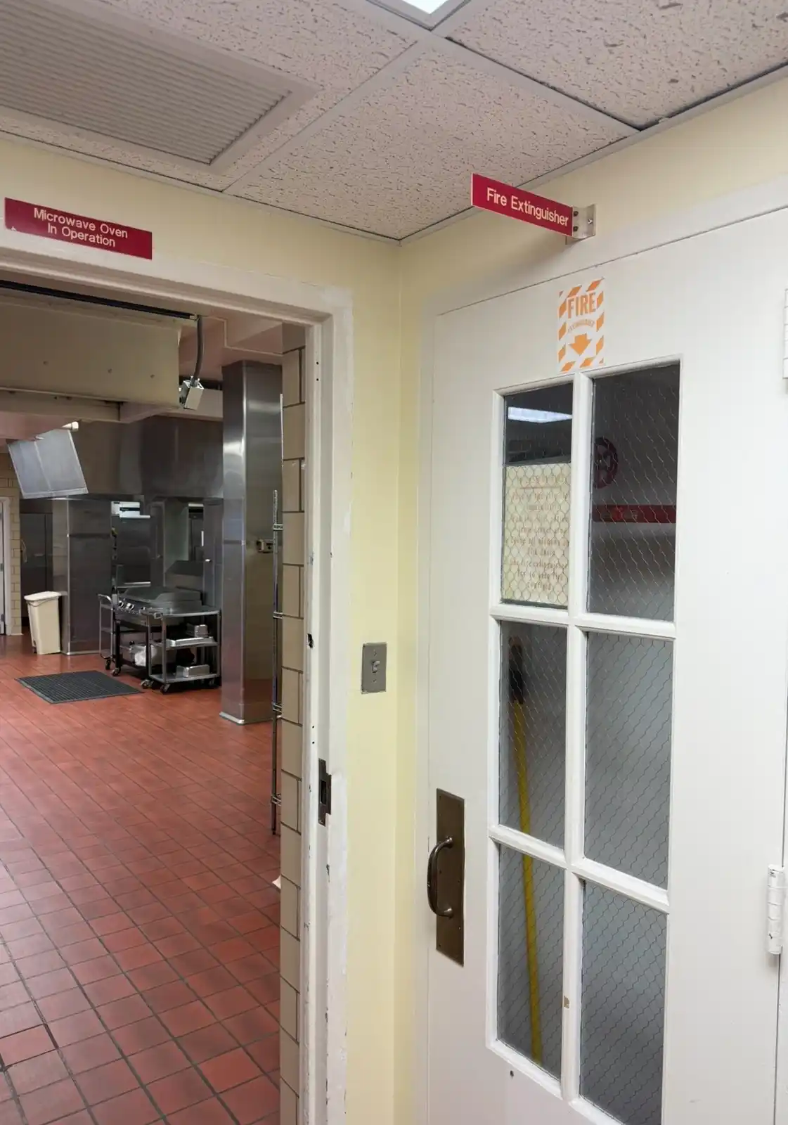 A photo of a commercial kitchen hallway with a red sign above a doorway reading 'Microwave Oven In Operation' and another…