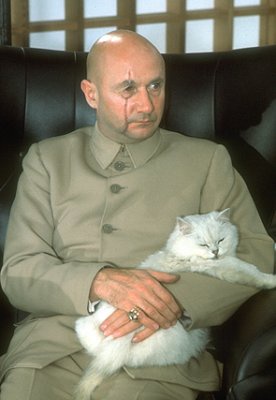 a bald man in a beige uniform sits in a leather chair holding a white fluffy cat. The cat is curled up and sleeping. The man…