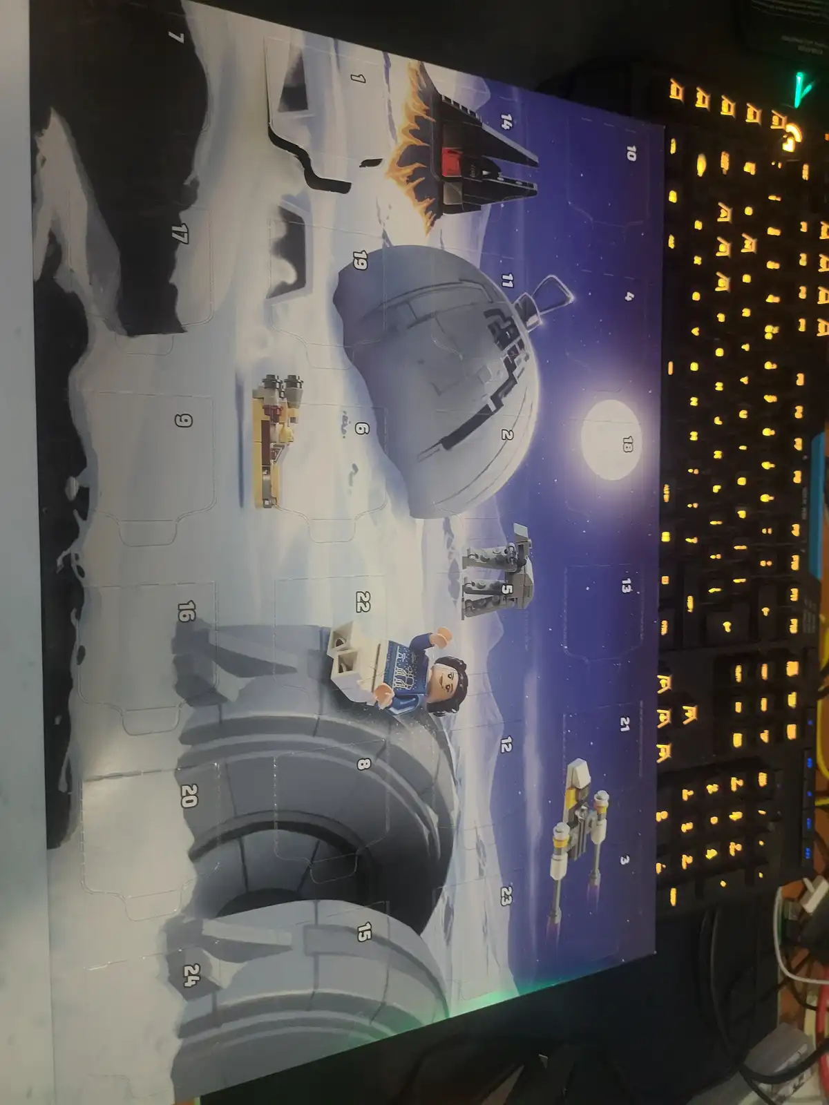 A Lego Star Wars Advent Calendar is laid out on a desk next to a lit-up keyboard, with the calendar's numbered compartments…